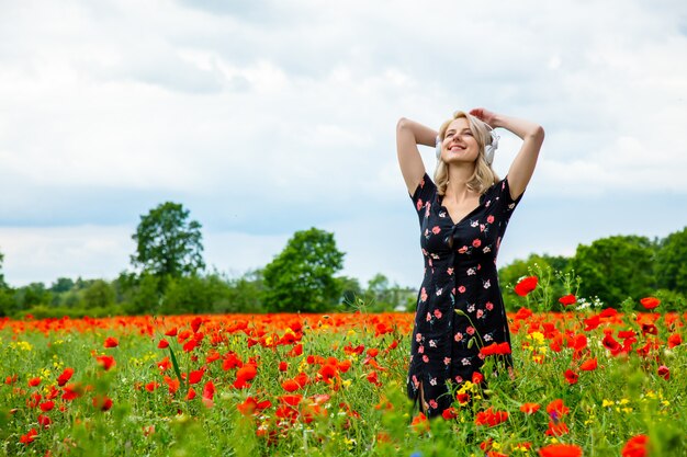 Chica rubia en un hermoso vestido con auriculares en el campo de amapolas en verano