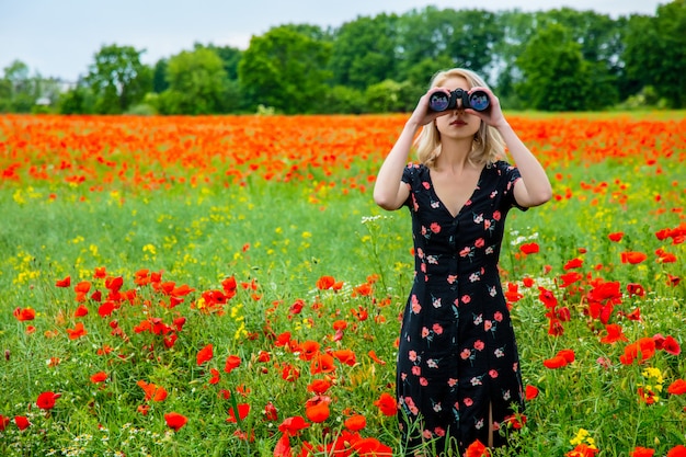 Chica rubia en un hermoso vestido con binoculares en el campo de amapolas en verano