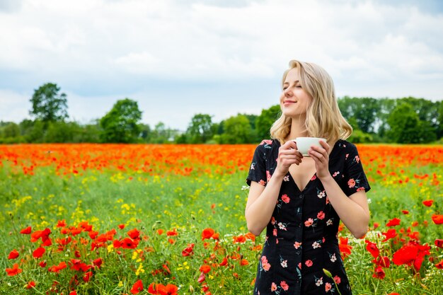 Chica rubia en un hermoso vestido con una taza de café en el campo de amapolas en verano