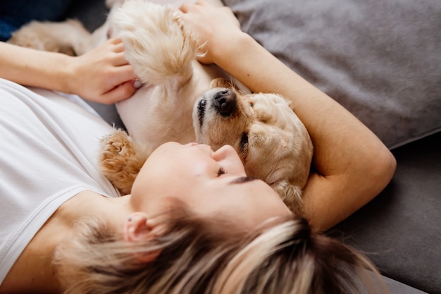 Chica rubia en jeans está acostada de espaldas en un sofá gris con un perro