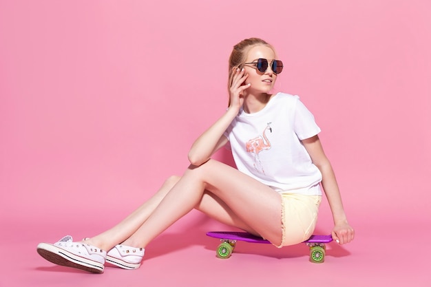 Chica rubia joven alegre con una patineta en el fondo de color rosa.