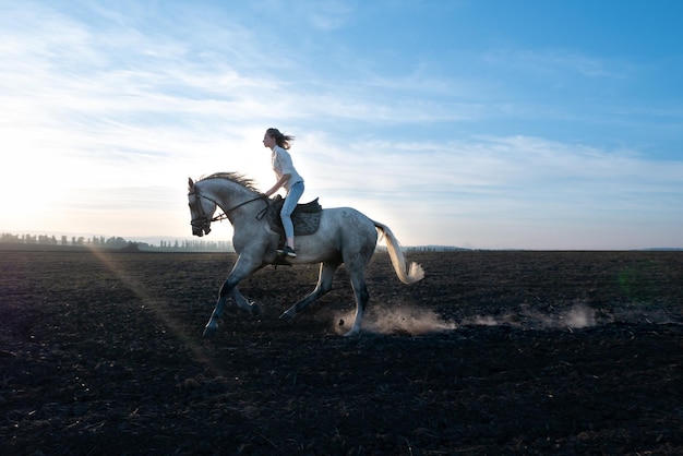 Chica rubia joven cabalgando sobre un caballo en el campo durante la puesta de sol. Los cascos de los caballos levantan polvo mientras corren sobre suelo seco