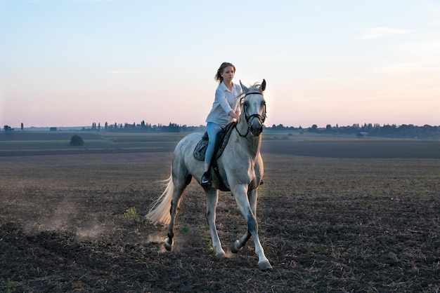 Chica rubia joven cabalgando sobre un caballo en el campo durante la puesta de sol. Los cascos de los caballos levantan polvo mientras corren sobre suelo seco