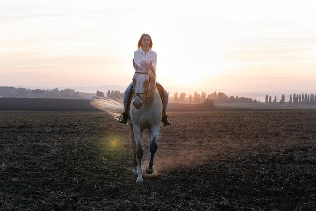 Chica rubia joven cabalgando sobre un caballo en el campo durante la puesta de sol