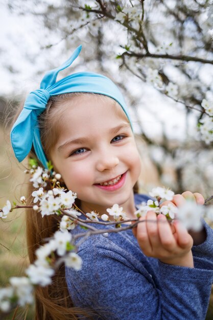 Chica rubia joven de pie en un jardín floreciente. Cereza floreciente. Retrato de niña hermosa. Cerca de la pequeña cara de modelo. Sakura floreciendo, tarde de primavera