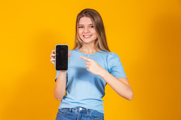 Chica rubia joven que muestra la pantalla del teléfono inteligente en blanco y apunta mientras mira a la cámara y sonríe.