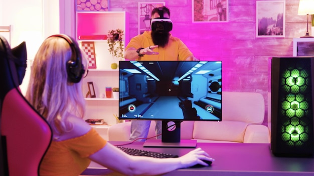 Chica rubia jugando juegos de disparos en línea sentado en una silla de juego. Novio con gafas de realidad virtual.