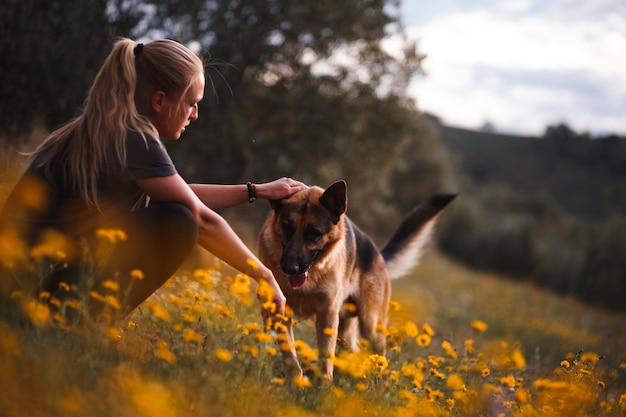 Chica rubia jugando con perro pastor alemán en un campo de flores amarillas