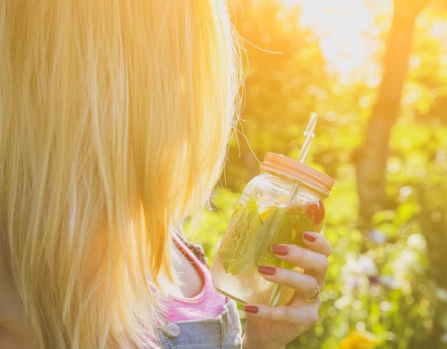 Chica rubia con limonada fresca en tarro con paja. Bebidas de verano hipster. Estilo de vida vegano saludable. Ecológico en la naturaleza. Limones, naranjas y bayas con menta en el vaso.