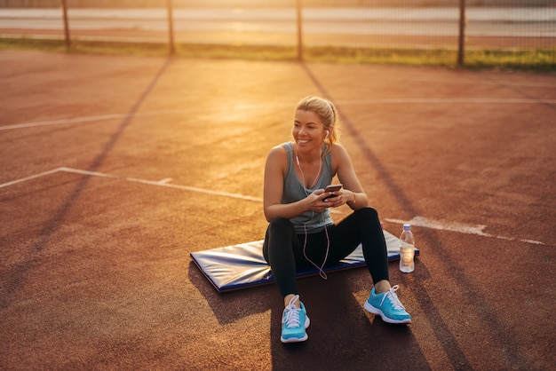 Chica rubia linda deportiva joven cansada que se sienta afuera en una estera de entrenamiento después de entrenar duro y escuchar la música.