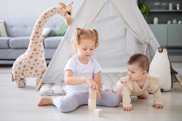 Una chica rubia en una luminosa habitación infantil juega cubos de madera con su hermana menor en el wigwam y se regocija y sonríe