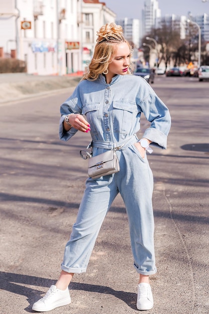 Chica rubia con maquillaje brillante y peinado en traje de mezclilla posando en una calle de la ciudad