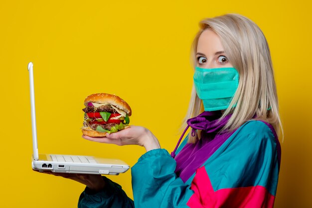 Chica rubia en mascarilla con hamburguesa y computadora portátil