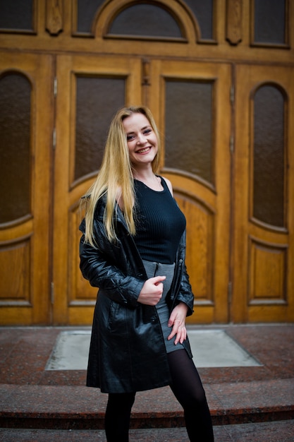 Chica rubia de moda en largo abrigo de cuero negro posado contra la puerta de madera.