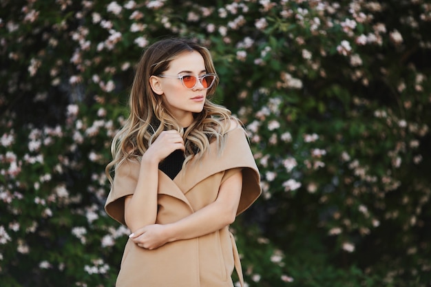 Chica rubia modelo con abrigo sin mangas y elegantes gafas de sol posando al aire libre