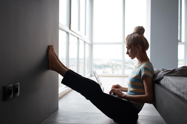 Chica rubia con moño trabajando en casa escribiendo en una laptop sentada en el suelo