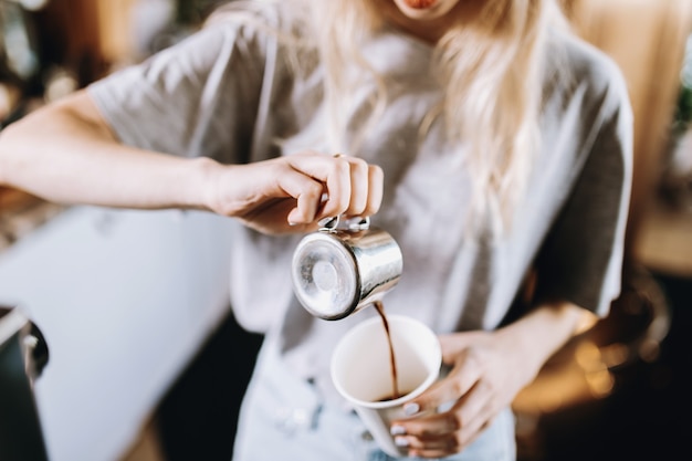 Una chica rubia muy delgada con cabello largo, vestida con ropa casual, vierte café en un vaso en una acogedora cafetería. .