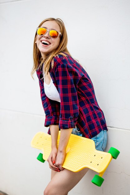 una chica rubia muy sonriente está de pie frente a la pared gris y sostiene un longboard amarillo