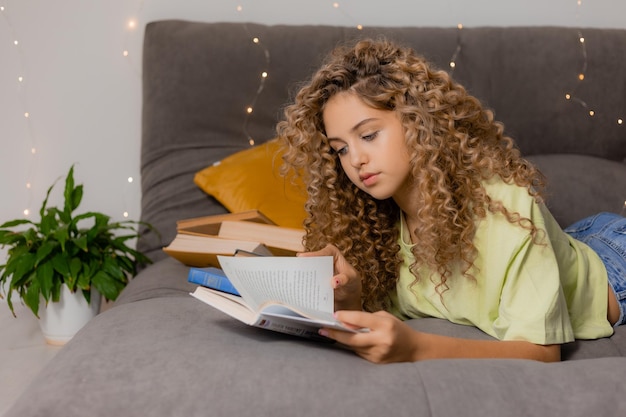 Chica rubia con ojos azules y cabello rizado está leyendo un libro acostado en una cama gris