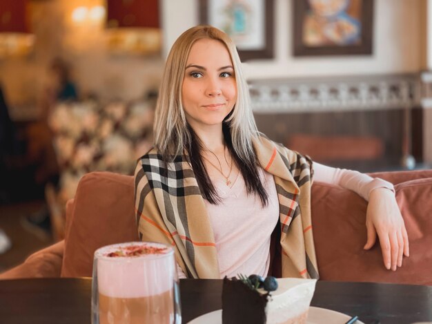 Una chica rubia con un pañuelo en los hombros con un suéter rosa suave se sienta en un restaurante acogedor y está relajada y toma un café y un pedazo de pastel en su mesa.