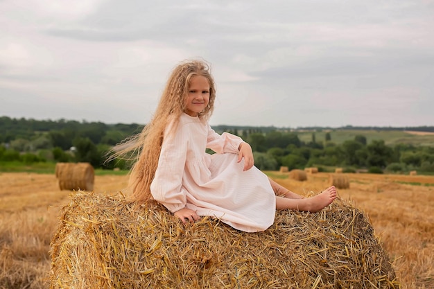 una chica rubia con el pelo largo y un vestido de lino rosa está sentada en un pajar en un campo segado