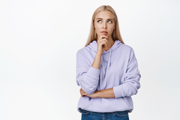 Chica rubia pensativa que busca una solución tocando la barbilla mientras reflexiona sobre la decisión mirando a la esquina superior izquierda de pie sobre fondo blanco