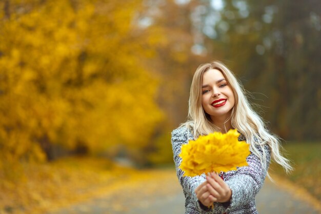 Chica rubia positiva con labios rojos con elegante abrigo con hojas amarillas en el bosque de otoño. Espacio para texto