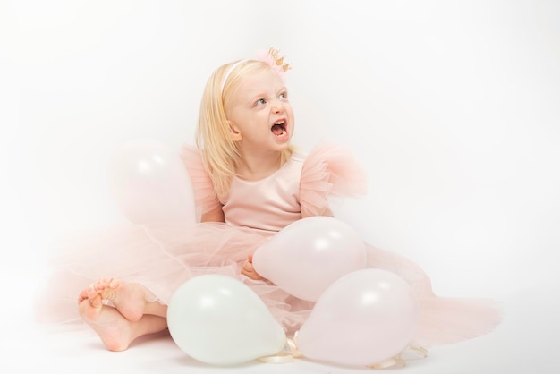 Chica rubia riendo en vestido rosa exuberante con globos se sienta en el piso Niño feliz en su cumpleaños Fondo blanco