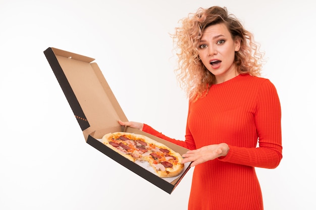 Chica rubia rizada sorprendida en un vestido rojo tiene una caja de pizza en blanco