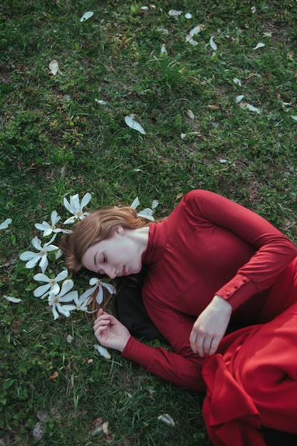 Una chica rubia de rojo yace en la primavera sobre la hierba rodeada de flores de magnolia