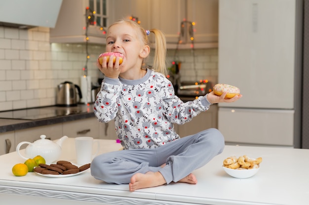 Chica rubia en ropa estampada de Navidad está sentada en la hermosa cocina con dos donas en sus manos