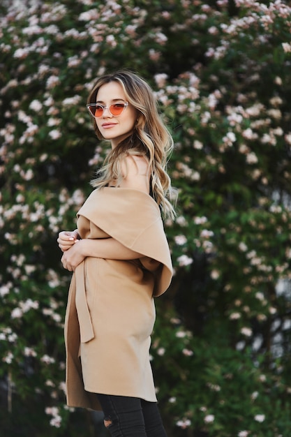 Chica rubia sensual y de moda con abrigo sin mangas y elegantes gafas de sol