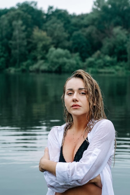 Chica rubia sensual con el pelo mojado en una camisa blanca y traje de baño negro posando en el lago.