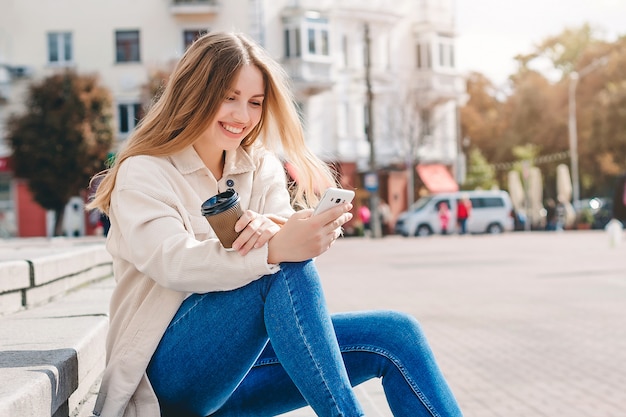 Chica rubia sentada en los escalones con un teléfono móvil riendo y enviando mensajes de texto con el mensajero