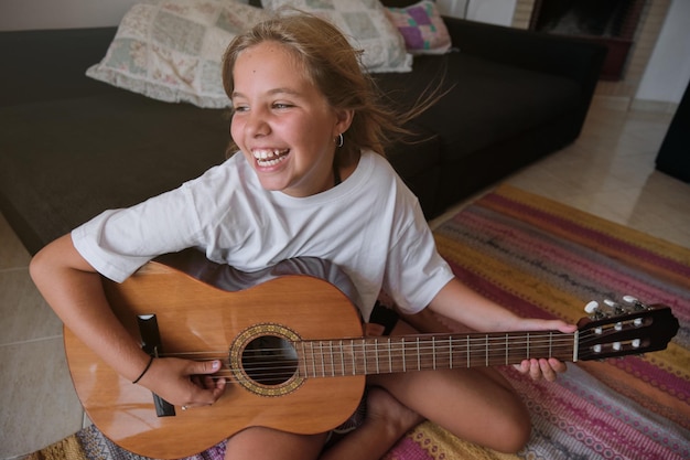 Una chica rubia sentada en el suelo de una casa tocando una guitarra mientras canta con expresión feliz y mirando a un lado