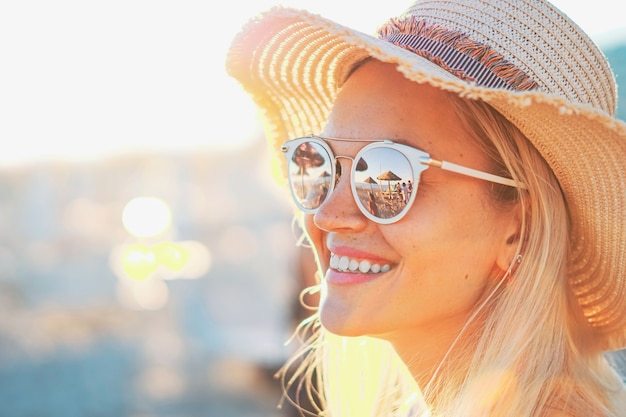 Una chica rubia sonriendo con sombrero - Playa y luz del sol reflejada en gafas de sol - Hermosas mujeres jóvenes en vacaciones - Foto de retrato