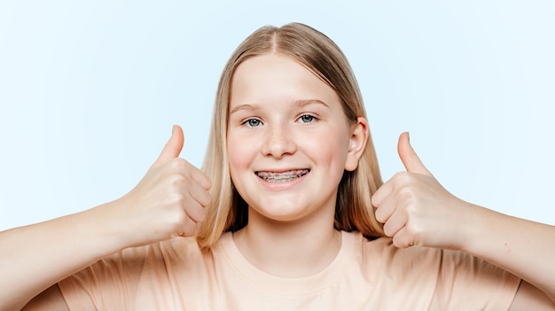 Chica rubia sonriente con aparatos de ortodoncia sosteniendo sus pulgares hacia arriba, tratamiento de ortodoncia