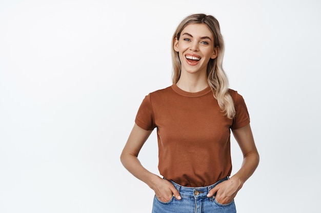 La chica rubia sonriente se ve entusiasmada con la cámara y usa una camiseta informal con jeans en una pose relajada contra el fondo blanco