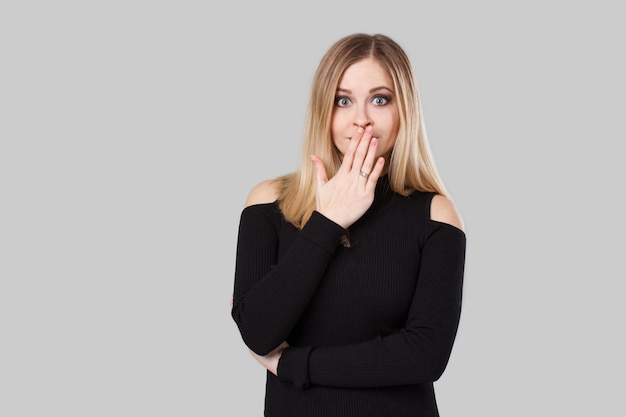 La chica rubia se sorprende y se cubre la boca con la mano. Ella de pie frente a un fondo gris. Lleva un suéter negro.