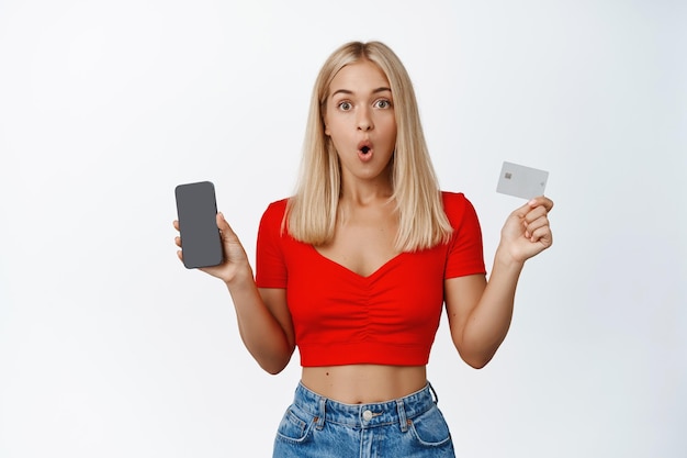 Chica rubia sorprendida muestra la tarjeta de crédito de la pantalla del teléfono inteligente en la mano hablando de la aplicación de compras de teléfonos móviles recomienda la aplicación de fondo blanco