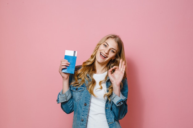 Una chica rubia sosteniendo un pasaporte con boletos de viaje y sonriendo sobre un fondo rosa