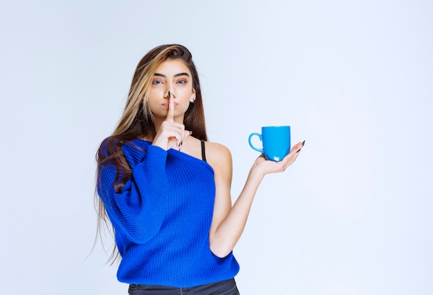 Chica rubia sosteniendo una taza de café azul y pidiendo silencio.