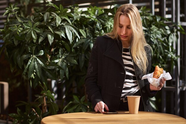 Chica rubia sosteniendo una taza de papel de café y muffin sentado en un café. concepto de comida callejera