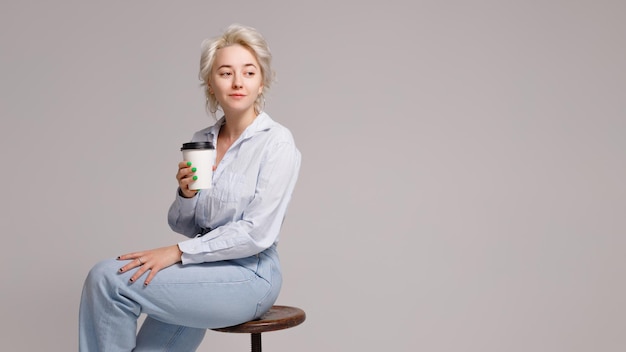 Chica rubia sosteniendo un vaso de plástico con una bebida en sus manos sobre un fondo claro. Foto de alta calidad