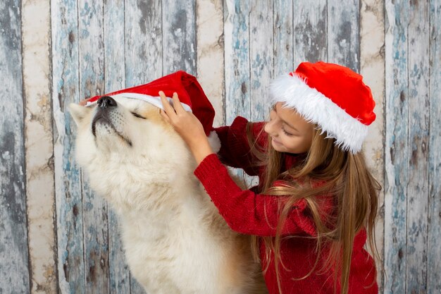 Chica rubia con su perro con gorro de Papá Noel, poniendo el sombrero en su perro, celebrando la pared de madera de Navidad