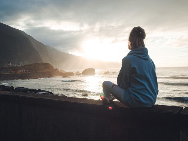 Foto chica rubia con sudadera con capucha, sentada cerca del océano disfrutando de la vista durante el atardecer