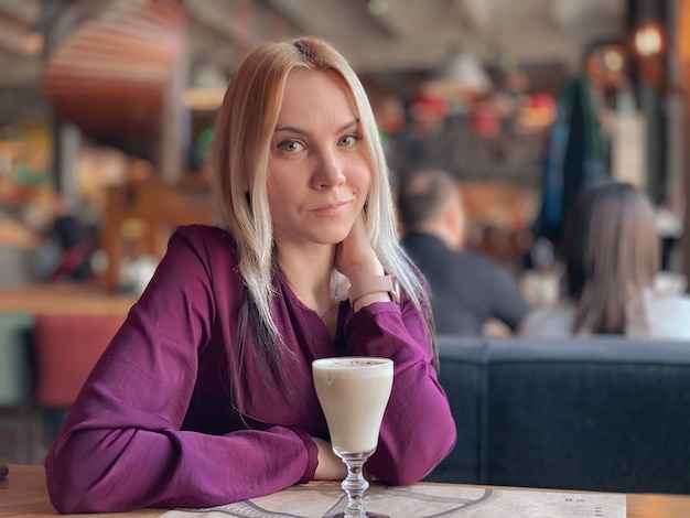 Una chica rubia con un suéter lila se sienta en un sofá en un acogedor restaurante y ella está relajada y sonriente y toma un café en su mesa.