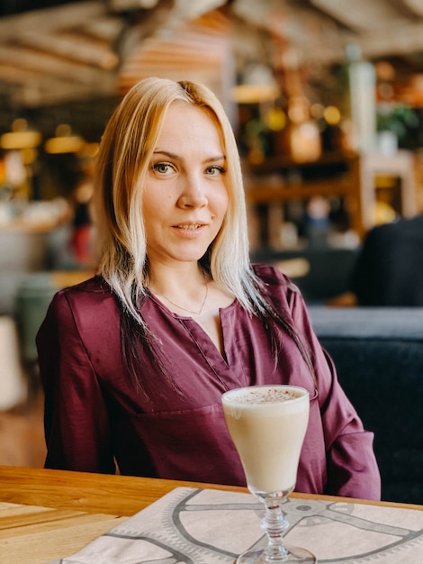 Una chica rubia con un suéter lila se sienta en un sofá en un acogedor restaurante y ella está relajada y sonriente y toma un café en su mesa.