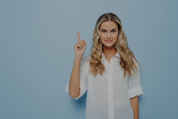 Chica rubia tranquila con una leve sonrisa apuntando hacia arriba en el aire con su dedo, tiene una nueva idea brillante y la muestra con un gesto mientras está de pie junto a la pared azul. Mujer mostrando en el espacio de la copia