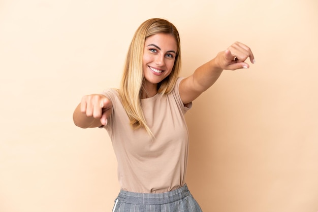 Chica rubia uruguaya aislada sobre fondo beige apuntando al frente con expresión feliz